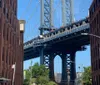 This image features pedestrians and vehicles on a bustling street framed by old brick buildings with the iconic steel structure of the Manhattan Bridge towering in the background under a clear blue sky