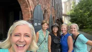 A group of five smiling women takes a selfie together in front of a historic brick building with an arched metal door.