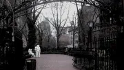 A grayscale image showing a serene park pathway with benches, leafless trees, a street lamp, and sculptures depicting people in an urban setting.