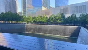 The image shows one of the 9/11 Memorial pools with names inscribed around it, reflecting the surrounding high-rise buildings and trees under a clear blue sky.