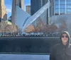 A person in sunglasses and a jacket is standing in front of the 911 Memorial with the distinctive white wing-like structure of the Oculus in the background