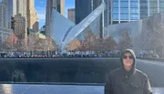 A person in sunglasses and a jacket is standing in front of the 9/11 Memorial with the distinctive white, wing-like structure of the Oculus in the background.