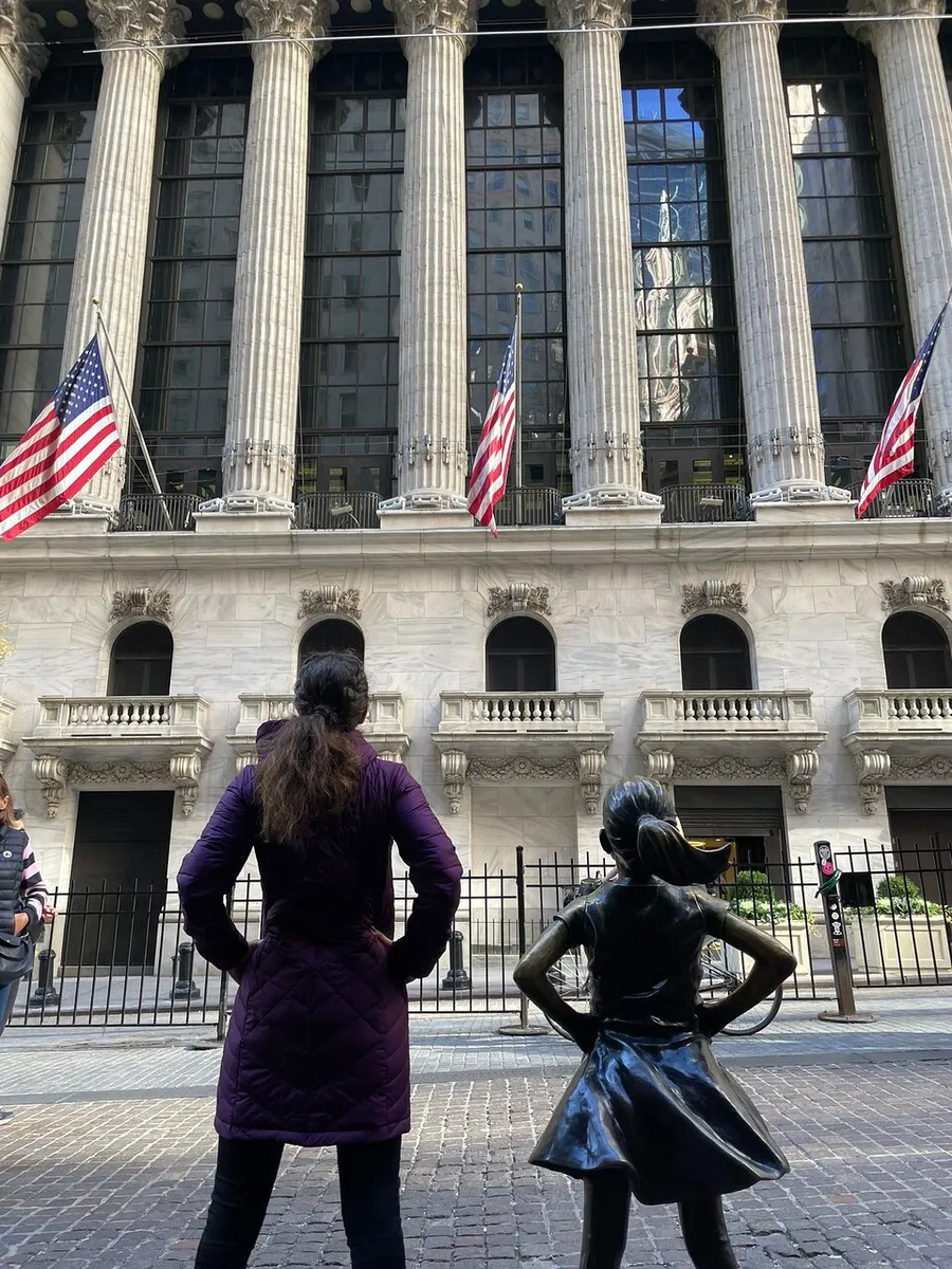 A woman stands next to the Fearless Girl statue, both facing the New York Stock Exchange building adorned with American flags.