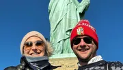 Two people are smiling for a selfie with the Statue of Liberty in the background.