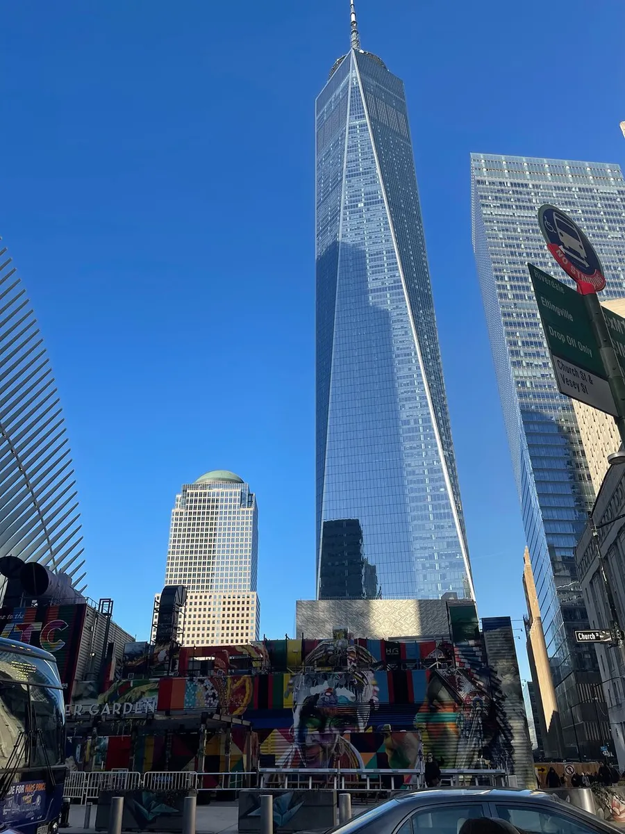 The image shows a bustling street scene with the towering One World Trade Center in the background, under a clear blue sky.
