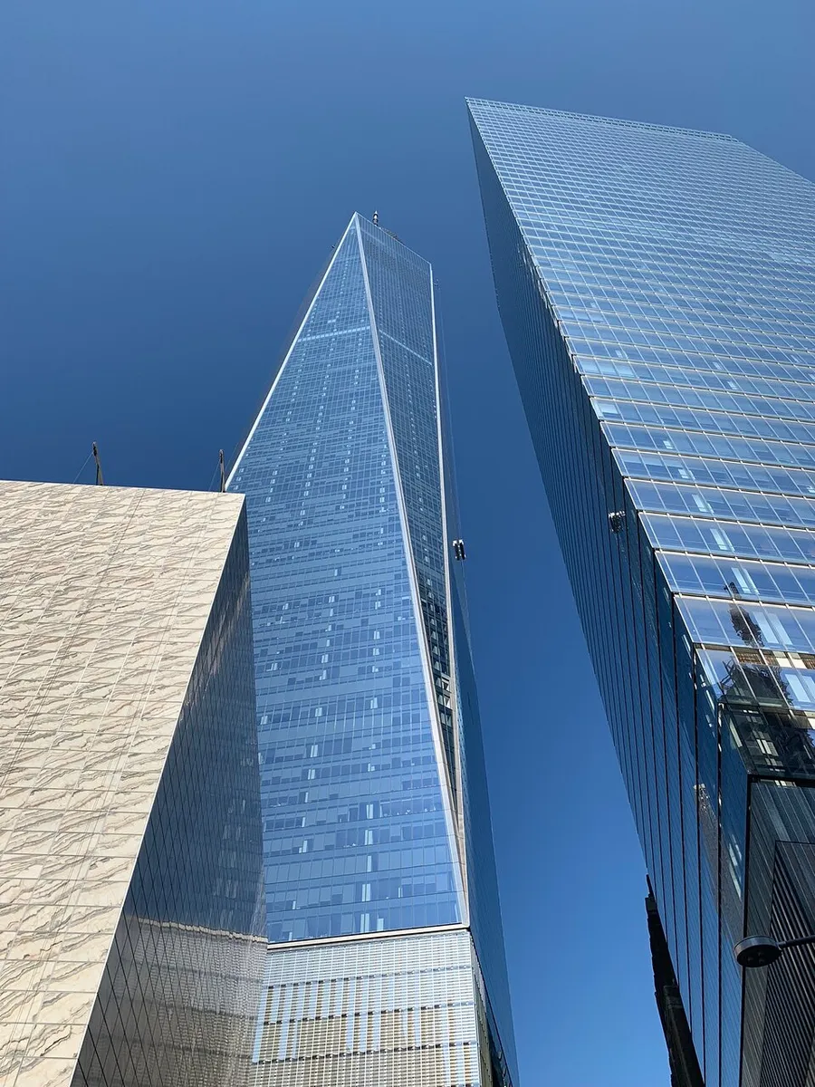 The image shows a worm's-eye view of tall skyscrapers against a clear blue sky, with one building reflecting the sky on its glass facade.