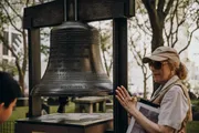 A woman, wearing a cap and sunglasses, admires a large bell mounted on a frame that bears an inscription, with a partial view of a young person on the left.