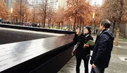 Two individuals are standing beside a reflective memorial with names inscribed on it, as the woman points upwards towards the surrounding urban landscape or sky, amidst a setting of bare trees and overcast weather.