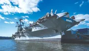 The image shows the aircraft carrier USS Intrepid, now a sea, air, and space museum, docked at a harbor under a blue sky with scattered clouds.