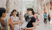 A tour guide is enthusiastically speaking to visitors inside a sunlit museum gallery adorned with classical sculptures.