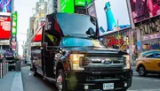 A black Ford truck with its hood open is parked on a busy street in Times Square, amid bright digital billboards and passing traffic, including a yellow cab.