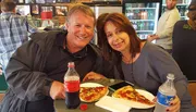 A smiling man and woman are enjoying slices of pizza and soft drinks while sitting at a table.