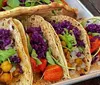 The image shows a colorful platter of vegetarian tacos filled with purple cabbage greens roasted vegetables and chickpeas accompanied by a green sauce