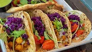 The image shows a colorful platter of vegetarian tacos filled with purple cabbage, greens, roasted vegetables, and chickpeas, accompanied by a green sauce.
