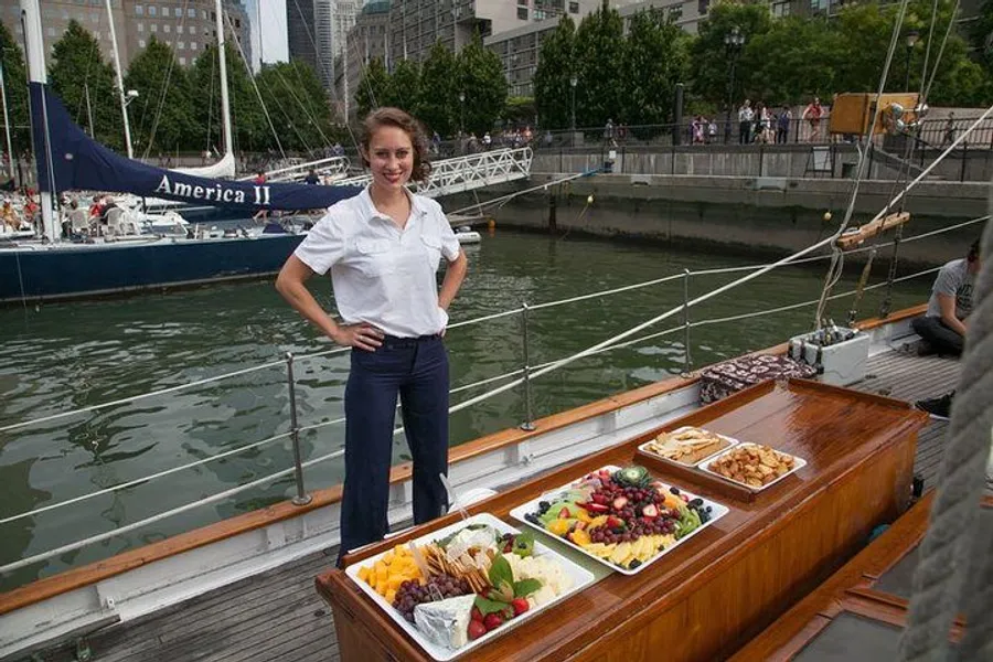 A woman stands smiling on a boat next to a buffet spread of fruits, cheeses, and other snacks, with another boat and a waterfront promenade in the background.