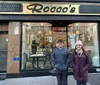 A young man and woman are smiling in front of Pasticceria Rocco a bakery with a cozy interior visible through its glass facade