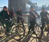A group of smiling people wearing helmets are standing with bicycles on a sunny boardwalk by the water with a bridge in the background
