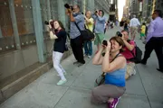Several individuals are photographing something off-camera on a city sidewalk, some using unique stances to capture their shot.