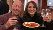 A smiling man and woman are holding glasses of red and white wine, respectively, with a plate of what appears to be cannoli between them, sharing a cheerful moment.