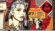 The image shows a colorful urban mural featuring a graphic representation of the iconic lead singer of the band Blondie, flanked by concert posters, with three individuals posing in front of it.