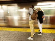 Two people stand on a subway platform as a blur of a train passing by is captured in the background.