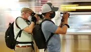 Two individuals are capturing a photograph or video, presumably of a moving train, as they stand beside a subway platform.