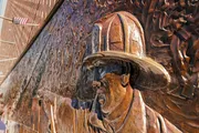 This image features a close-up view of a firefighter's face in profile, sculpted in relief on a bronze memorial wall, with a blurred American flag flying in the background.