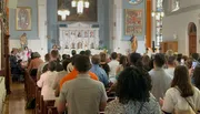 The image depicts a congregation of people standing and facing the altar inside a church, with several individuals capturing the moment on their phones.