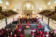 The image depicts a church congregation, with many individuals dressed in red garments, during a service with Christmas decorations adorning the interior.