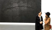 Two people are having a conversation in front of a large, textured black painting in an art gallery.
