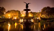 The image captures a beautifully lit angelic statue atop a fountain with water reflections in a serene park at twilight.