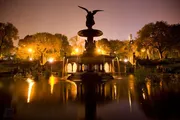 The image captures a nighttime view of the illuminated Bethesda Fountain in Central Park, New York City, with a reflective pool in the foreground and ambient city lights peering through the trees in the background.