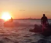 Two people are riding jet skis on a body of water with a sunset in the background and a city skyline is faintly visible in the distance