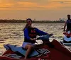 Two people are riding jet skis on a body of water with a sunset in the background and a city skyline is faintly visible in the distance