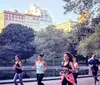 A group of people are jogging through a lush park with a skyline of tall buildings in the background suggesting a vibrant urban park environment