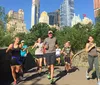 A group of people are jogging through a lush park with a skyline of tall buildings in the background suggesting a vibrant urban park environment