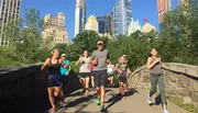 A group of people are jogging through a lush park with a skyline of tall buildings in the background, suggesting a vibrant urban park environment.