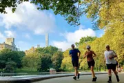 Three people are jogging in a park with a backdrop of skyscrapers and lush trees.