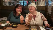 Two people are smiling and posing with chopsticks and food at a restaurant table.