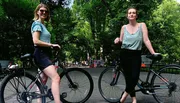 Two people are smiling and standing next to bicycles in a park with trees and pedestrians in the background.