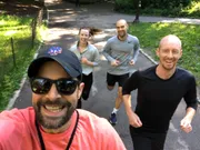 Four people, one taking a selfie, are smiling and jogging on a sunny day on a park path.