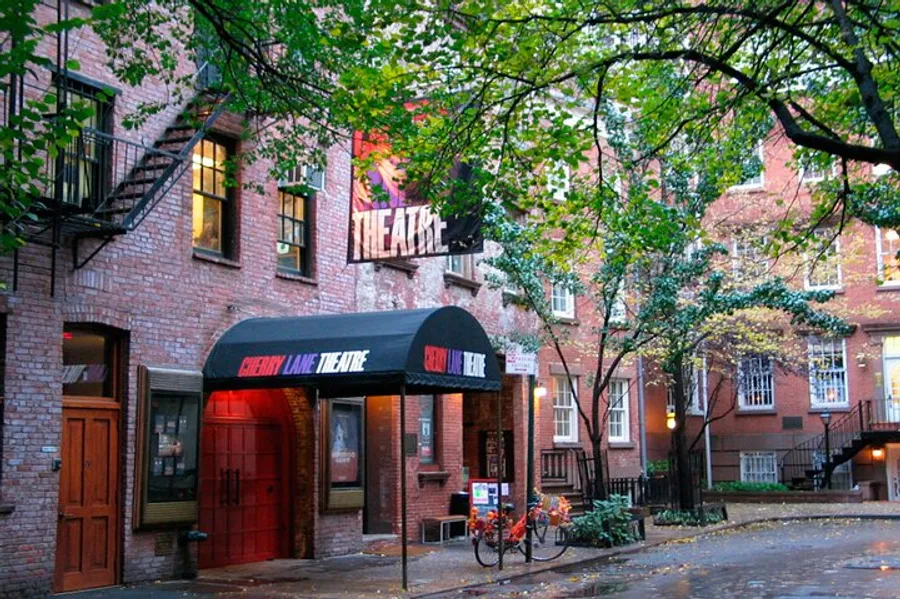 The image shows the Cherry Lane Theatre with its classic red brick facade and black awnings, tucked away on a leafy street that gives off a quaint and historic atmosphere.