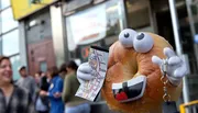 A personified bagel with eyes, mouth, and hands holds a map and appears to be a tourist, creating a playful and whimsical scene on an urban street.