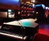 The image shows a dimly lit jazz club with a grand piano in the foreground and a bar with colorful shelves in the background