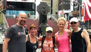 Five people are posing together for a photo in what appears to be Times Square, with a statue and the iconic red steps in the background.