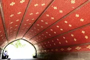 A person is sitting under a curved, red-patterned brick underpass or tunnel with daylight showing at the end.