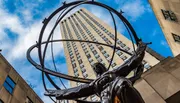 A statue of Atlas holding the celestial spheres stands before the art deco facade of a skyscraper, under a blue sky with scattered clouds.