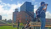 A person is taking a photograph while others enjoy a sunny day on a rooftop garden overlooking a mix of historic and modern urban architecture.