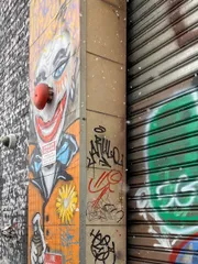 The image shows an urban wall with a vibrant clown graffiti artwork, alongside various tags and a partially visible metal shutter.