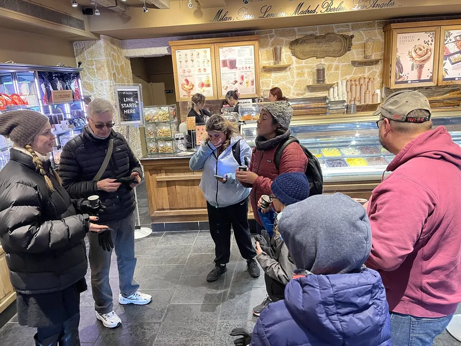 A group of people, dressed warmly, are waiting in line at a gelateria, with some engaged in conversation and others looking at their phones.